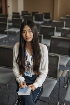 Asian woman in business attire sitting in an empty conference room, holding a notebook.