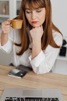 Focused woman in white shirt pondering at laptop with coffee cup and book.