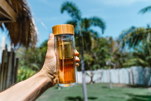 Hand holding a glass tea tumbler with bamboo lid, vibrant greenery background, West Nusa Tenggara.