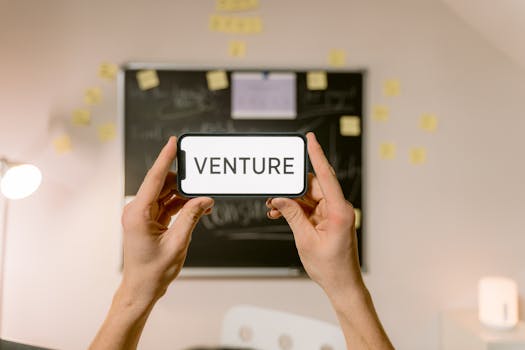 A smartphone displaying 'Venture' held by hands in an office setup. Ideal for business and startup themes.