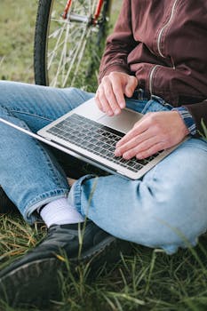 Close-up of a person sitting on grass, typing on a laptop with a bicycle nearby.