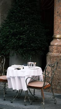 Charming outdoor café table set with two iron chairs and a white tablecloth in a courtyard.